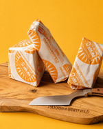 Three wrapped wedges of Paxton & Whitfield cheese arranged on a wooden cutting board with a matching branded knife, against a solid yellow background.