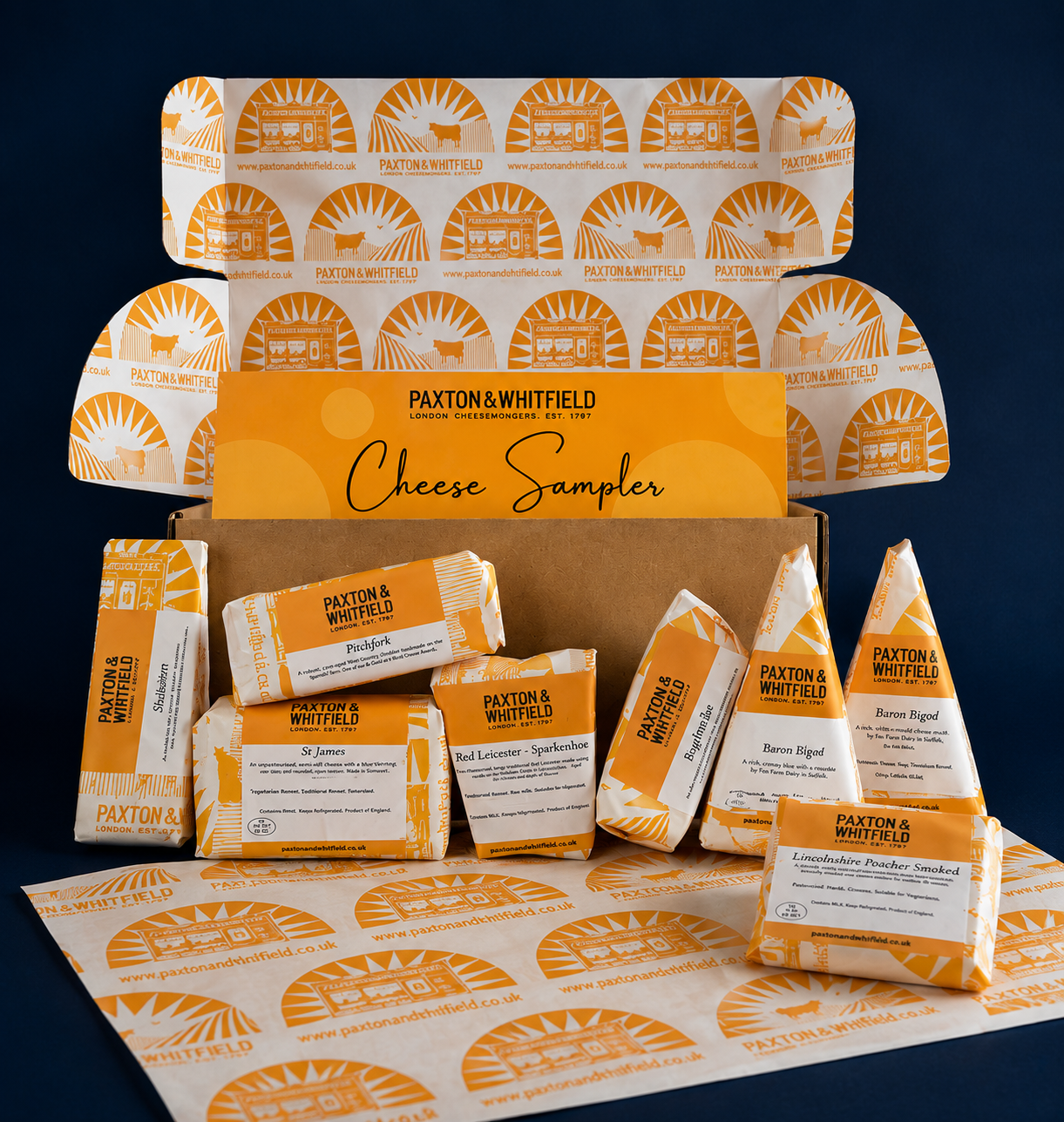 A curated cheese sampler gift set from Paxton & Whitfield, featuring multiple individually wrapped cheese wedges and blocks arranged around an open kraft box with branded orange-and-white paper packaging on a dark navy background.