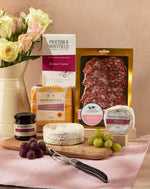 Assorted food items including a loaf of bread, salami, cheese, and grapes on a table with Paxton & Whitfield branding.